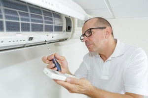 HVAC technician inspecting air conditioning unit with screwdriver and remote control, emphasizing maintenance for indoor air quality.