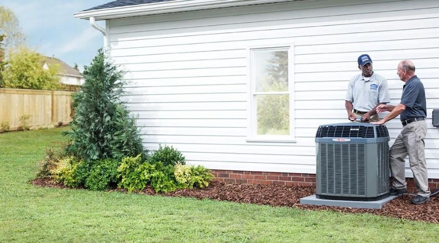 Two HVAC technicians near a white house with an air conditioning unit and well-maintained landscaping.