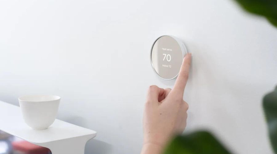 Smart thermostat being adjusted by a finger, showing 70 on a white wall, complemented by a green plant and simple bowl.
