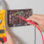 Hands holding testing probes with a multimeter and thermostat, showcasing HVAC maintenance and troubleshooting.